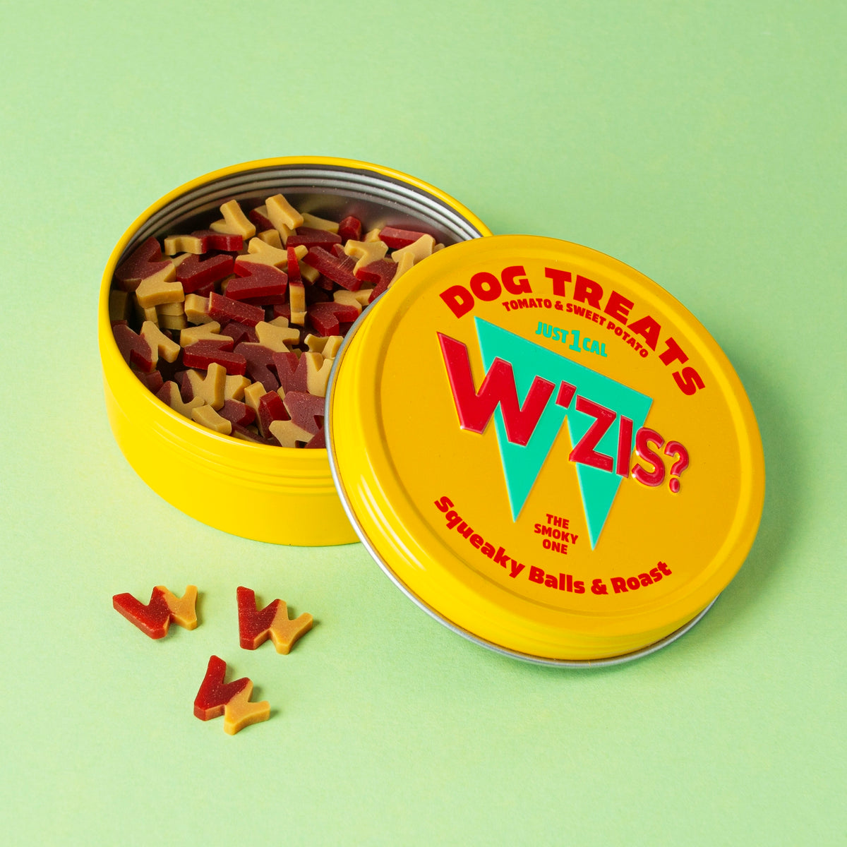 Yellow W'ZIS Squeaky Balls & Roast plant-based dog treat tin open beside colourful treats on a green background.