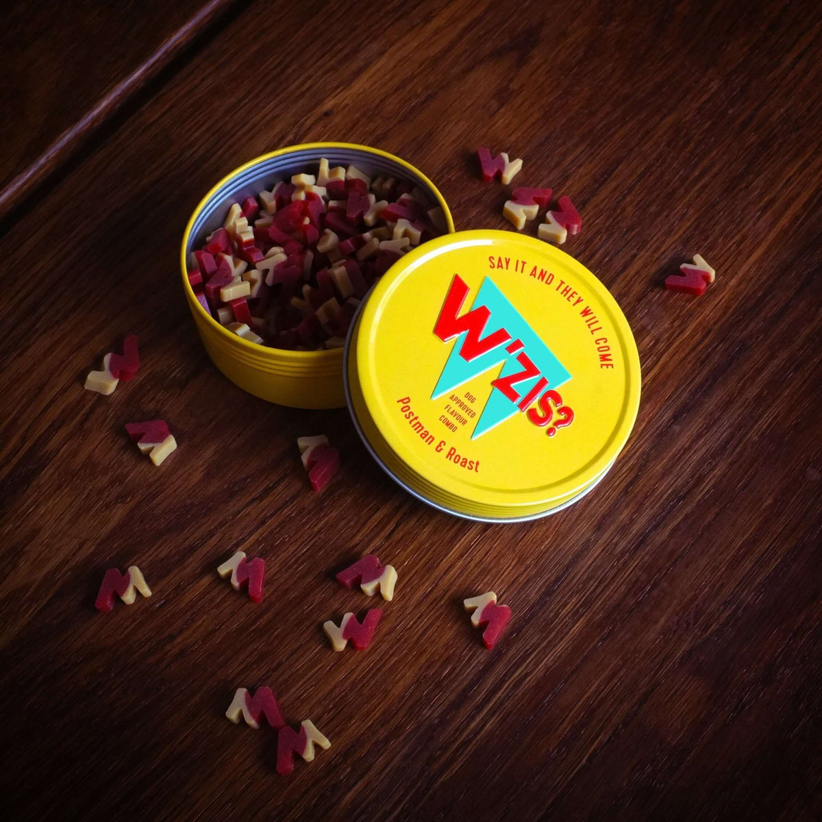 Open yellow W'ZIS Squeaky Balls & Roast treat tin with small plant-based dog treats scattered on a wooden table.